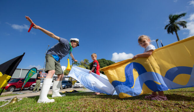 An outdoor Boats production photo. A Polyglot artist in shorts, t-shirt, white gumboots and a cap interacts with a small child who is holding a yellow and blue vessel around themselves. They are on green grass, and blue sky and a palm tree are visible in the background.