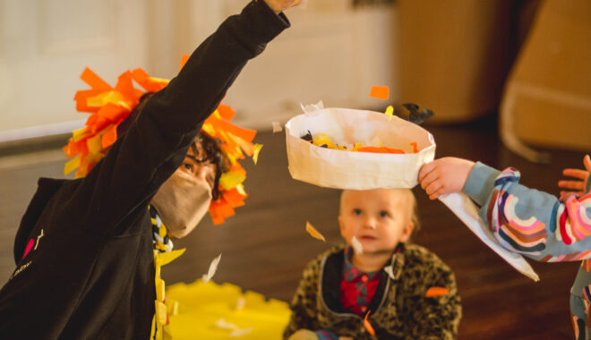 An indoor Paper Planet Production photo. A child looks up at an performer sprinkling streamer paper in a white paper basket. The streamer paper is yellow and orange. The performer is wearing a facemask, their right arm is lifted up and they are wearing a yellow and orange headpiece made out of streamers.