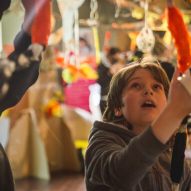 An indoor Paper Planet production photo. A child with a roll of masking tape on their hand is reaching up towards an orange tissue paper creation.