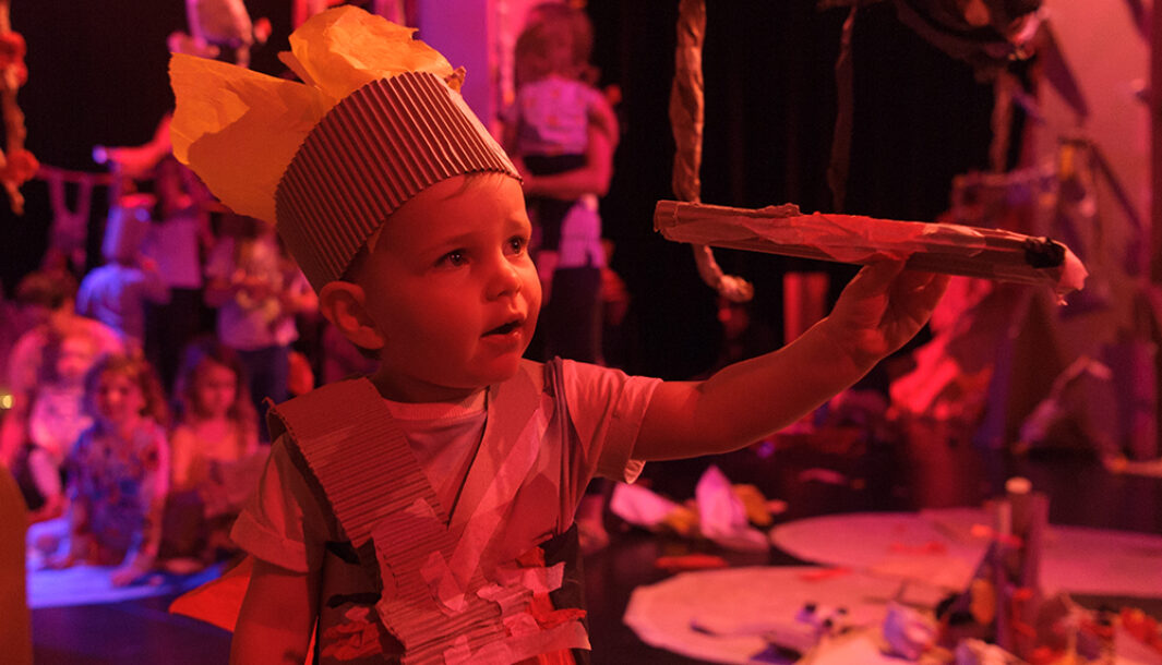 A Paper Planet production photo. A child wears a paper crown and armour made from cardboard. They hold a branch made from paper. They are surrounded by paper decorations, branches and vines made from paper. They are illuminated by soft pink light. Photo: Sarah Walker.