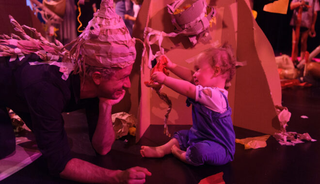 Baby and parent playing in creative paper world. An indoor Paper Planet production photo. A Polyglot artist in a paper costume leans on their elbows, gazing at a baby who is excitedly offering them some paper. They are next to a cardboard tree, and the scene is illuminated with pinkish theatre lighting.