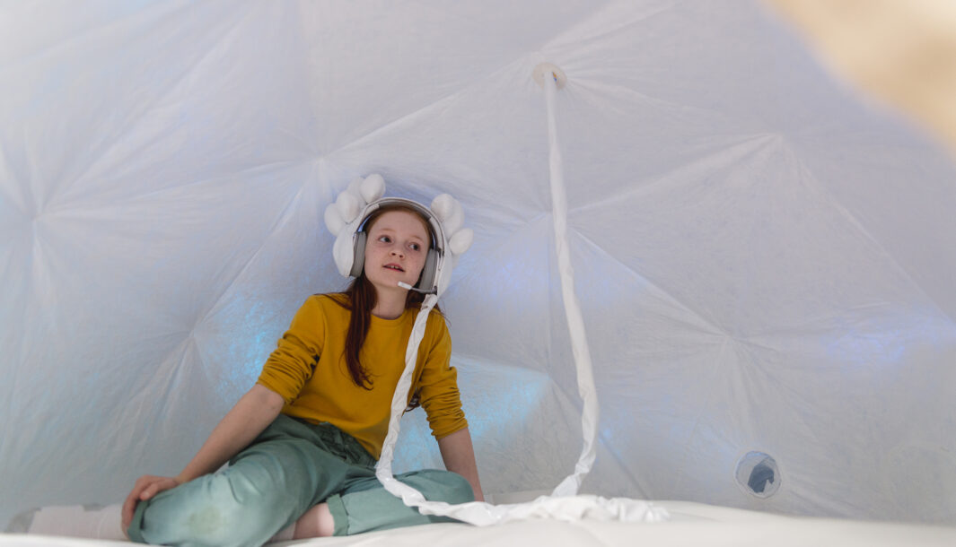Voice Lab production photo: Theresa Harrison. A child in a yellow jumper sits in a soft, white enclosed dome. They wear intricate, embellished white headphones that are attached to the space with a long cord.