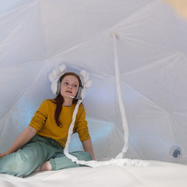 A child sits in an enclosed dome tent with headphones on. The headphones connect to a cable that runs out of the tent. The child sits on their side. The headphones are decorated with felt and baubles.