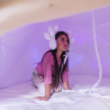 A Voice Lab production photo. A child kneels in a white dome, illuminated with soft purple light. They wear intricate white embellished headphones attached to the space with a long cord. Photo: Theresa Harrison.