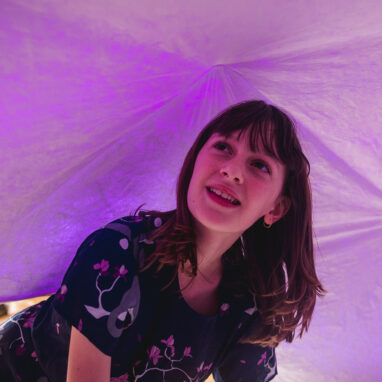A Voice Lab production image. A child with a fringe and shoulder-length hair crawls in a softly-lit dome tent. They are wearing a dress decorated with cherry-blossom patterns.