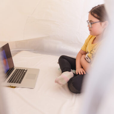 A Voice Lab production photo. A child sits in a softly-lit tent, wearing glasses and their hair tied back sits cross-legged and is viewing a laptop screen. The opening of the tent obscures the view of the child's body.