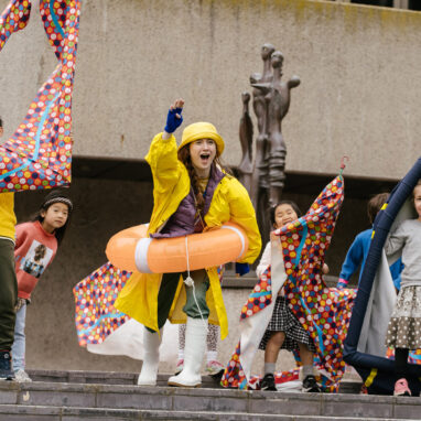 An outdoor Boats production photo. A Polyglot artist in a yellow raincoat, rain hat, white gumboots and orange flotation ring is shouting and gesturing excitedly from a set of stairs next to a statue. They are surrounded by excited, shouting children holding colourful vessels.