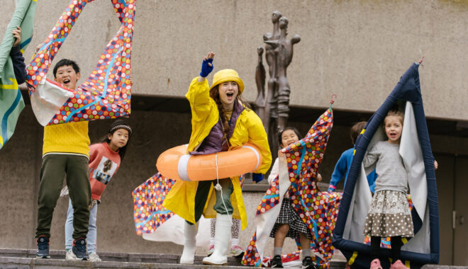 An outdoor Boats production photo. A Polyglot artist in a yellow raincoat, rain hat, white gumboots and orange flotation ring is shouting and gesturing excitedly from a set of stairs next to a statue. They are surrounded by excited, shouting children holding colourful vessels.