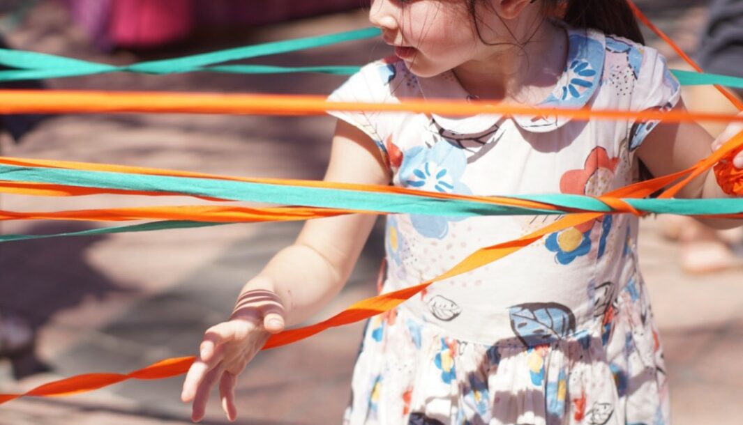 A Tangle production photo. A child looks sideways as they maneuver themselves amongst many coloured elastic strands. Some strands are in front of the child and some strands are behind the child.