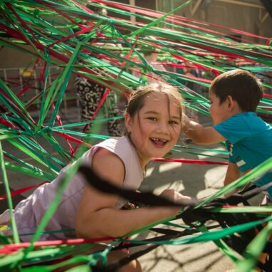 A Tangle production photo. A child smiles amongst elastic string surrounding them in different colours. Photo: Theresa Harrison.