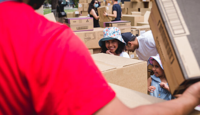 A We Built This City Production Photo. A child wearing a hat smiles and holds their hand to their face. Next to them, a smaller child smiles gleefully. The taller child's body is obscured by a large cardboard box. The top half of the smaller child's body is visible.