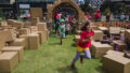 A We Built This City production photo. Children run excitedly through the elaborate cardboard entrance into the performance area, which is green lawn filled with brown cardboard boxes.