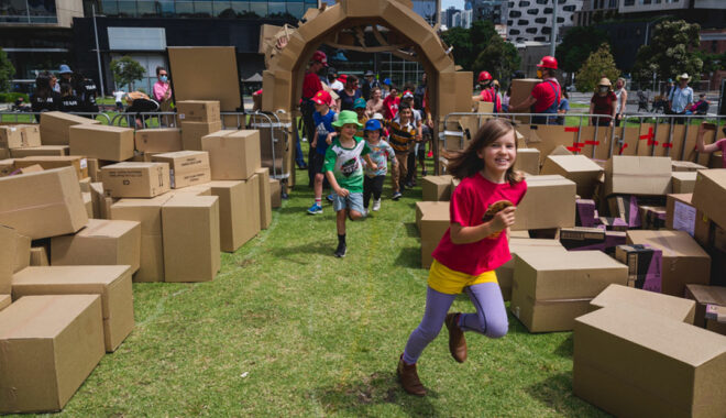A We Built This City production photo. Children run excitedly through the elaborate cardboard entrance into the performance area, which is green lawn filled with brown cardboard boxes.