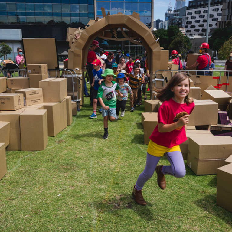 A We Built This City production photo. Children run excitedly through the elaborate cardboard entrance into the performance area, which is green lawn filled with brown cardboard boxes.