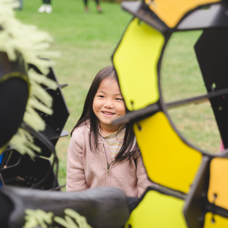An outdoor Bees production photo. A child with long dark hair in a pink jumper smiles up at a Polyglot artist who wears an intricate black and yellow Bee costume. They are standing next to the yellow and orange 'hive' set on a green lawn.
