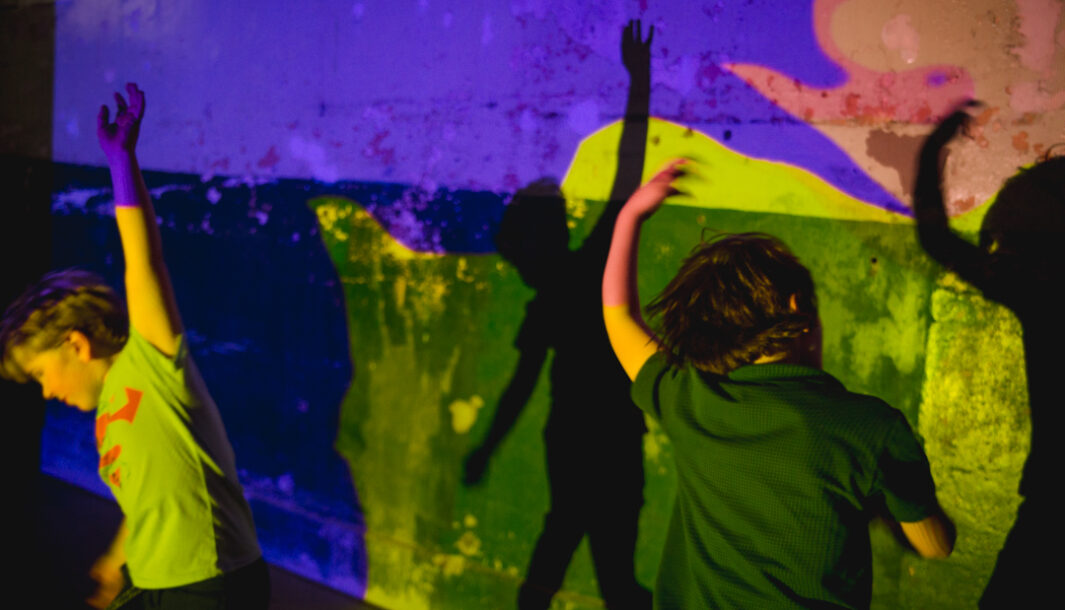 A Sound Shadows production photo. Two children dance in a darkened space, in front of a wall that is illuminated with brightly coloured projections, engaging with their shadows.
