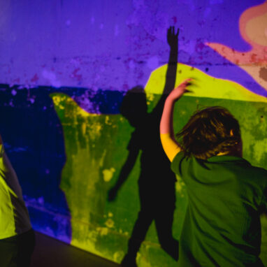 A Sound Shadows production photo. Two children dance in a darkened space, in front of a wall that is illuminated with brightly coloured projections, engaging with their shadows.