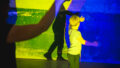 A Sound Shadows production photo. A child and an adult stand in a darkened space, engaging with their shadows on a wall, which is illuminated with bright yellow and blue projections.
