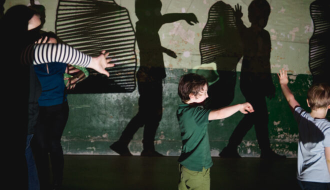 A Sound Shadows production photo. A group of adults and children move in a darkened space, illuminated with bright projections. Their shadows on the wall are augmented with abstract shapes.