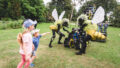 An outdoor Bees production photo. Two Polyglot artists in intricate black and yellow bee costumes engage with the blue, yellow and black 'hive' set piece. Two children watch, one pointing with their arm outstretched. Photographer: Theresa Harrison