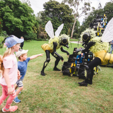 An outdoor Bees production photo. Two Polyglot artists in intricate black and yellow bee costumes engage with the blue, yellow and black 'hive' set piece. Two children watch, one pointing with their arm outstretched. Photographer: Theresa Harrison
