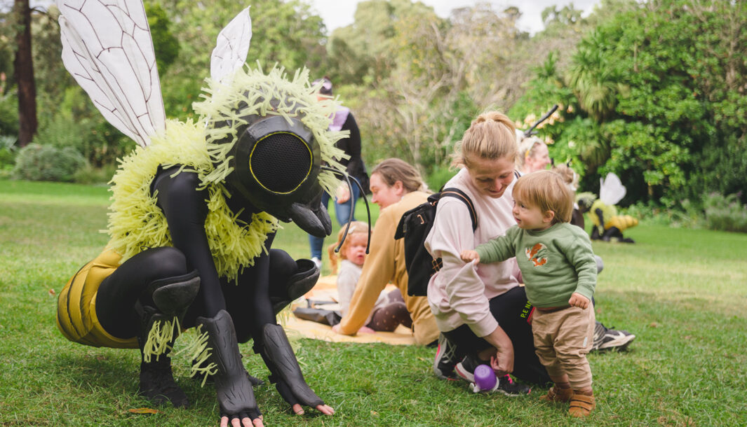 An outdoor Bees production photo. A Polyglot artist in an intricate black and yellow bee costume crouches on a green lawn. A small child, holding their parent's arm, looks at them intently. Other families are visible in the background, and the grassy area is surrounded by trees. Photographer: Theresa Harrison