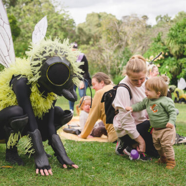 An outdoor Bees production photo. A Polyglot artist in an intricate black and yellow bee costume crouches on a green lawn. A small child, holding their parent's arm, looks at them intently. Other families are visible in the background, and the grassy area is surrounded by trees. Photographer: Theresa Harrison