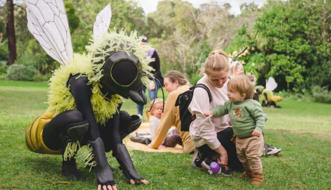 An outdoor Bees production photo. A Polyglot artist in an intricate black and yellow bee costume crouches on a green lawn. A small child, holding their parent's arm, looks at them intently. Other families are visible in the background, and the grassy area is surrounded by trees. Photographer: Theresa Harrison