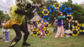 An outdoor Bees production photo. A Polyglot artist in an intricate black and yellow bee costume dances in front of the blue, yellow and black 'hive' set piece on a green lawn. They are surrounded by children, some wearing handmade paper bee costumes, following their movement. Trees are visible in the background.
