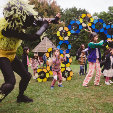 IMAGE: An outdoor Bees production photo. A Polyglot artist in an intricate black and yellow bee costume dances in front of the blue, yellow and black 'hive' set piece on a green lawn. They are surrounded by children, some wearing handmade paper bee costumes, following their movement. Trees are visible in the background. Photo by Theresa Harrison.