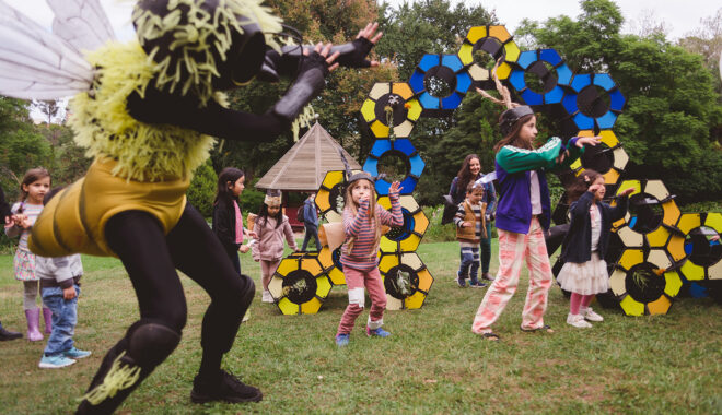 An outdoor Bees production photo. A Polyglot artist in an intricate black and yellow bee costume dances in front of the blue, yellow and black 'hive' set piece on a green lawn. They are surrounded by children, some wearing handmade paper bee costumes, following their movement. Trees are visible in the background.