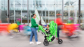 A Pram People photo. An adult in a green jumper and headphones pushes a small child in a pram decorated with green streamers. They are surrounded by colourful motion blurs. The adult looks out of frame, while the child looks at the camera. Photographer: Sarah Walker