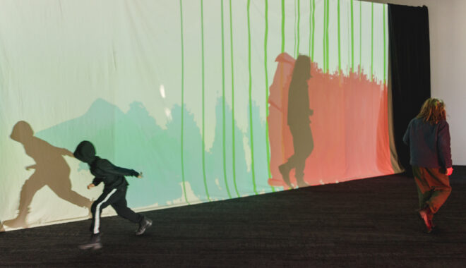 A Sound Shadows production photo. A child in a black hoodie and black pants crouches as he moves in front of a screen with colourful projections on it. Photographer: Theresa Harrison
