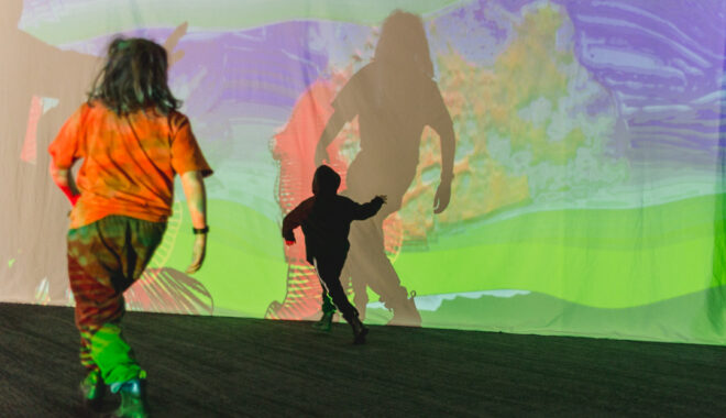 A Sound Shadows production photo. Two children play in front of a screen with colourful projections on it. One child is close up, the other is further away, playing with the shadow of the first child. Photographer: Theresa Harrison
