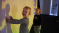 A Sound Shadows production photo. Two small children with short blonde hair stand in front of a large screen onto which colours are projected. One child is touching their shadow which falls on the screen, while looking at us and smiling. The other child stands close by and gazes up at the screen. Photographer: Theresa Harrison