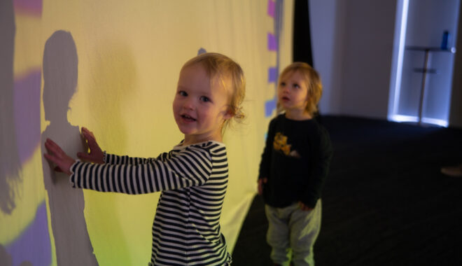 A Sound Shadows production photo. Two small children with short blonde hair stand in front of a large screen onto which colours are projected. One child is touching their shadow which falls on the screen, while looking at us and smiling. The other child stands close by and gazes up at the screen. Photographer: Theresa Harrison