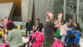 A Pram People production photo. A person claps their hands together. They have a pram decorated with streamers in front of them. Behind them, more parents talk with one another, also holding prams.