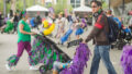 A Pram People production photo. A person pushes a pram and stares into the distance. The pram is decorated with streamers. Behind them, another participants walks in the opposite direction, pushing a decorated pram with streamers.