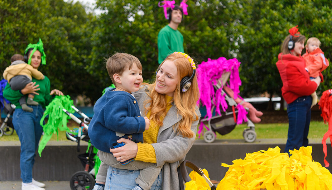 A Pram People photo. An adult in a yellow jumper, grey coat and headphones holds their child, smiling at them. Other adults with small children and brightly decorated prams stand in the background. They are outside on grey steps, with green trees in the background. Photographer: Sarah Walker