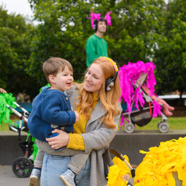 A Pram People photo. An adult in a yellow jumper, grey coat and headphones holds their child, smiling at them. Other adults with small children and brightly decorated prams stand in the background. They are outside on grey steps, with green trees in the background. Photographer: Sarah Walker