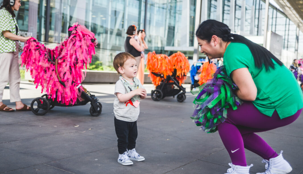 A Pram People production photo. A Polyglot artist in a colourful green and purple costume engages with a small child. Behind them are adults wearing headphones, standing with their prams decorated with brightly coloured ribbons. Photo: Theresa Harrison