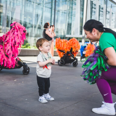 A Pram People production photo. A Polyglot artist in a colourful green and purple costume engages with a small child. Behind them are adults wearing headphones, standing with their prams decorated with brightly coloured ribbons. Photo: Theresa Harrison