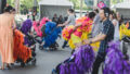 A Pram People production photo. People are walking adjacent to one another pushing black prams decorated with coloured streamers. All the participants are wearing headphones.