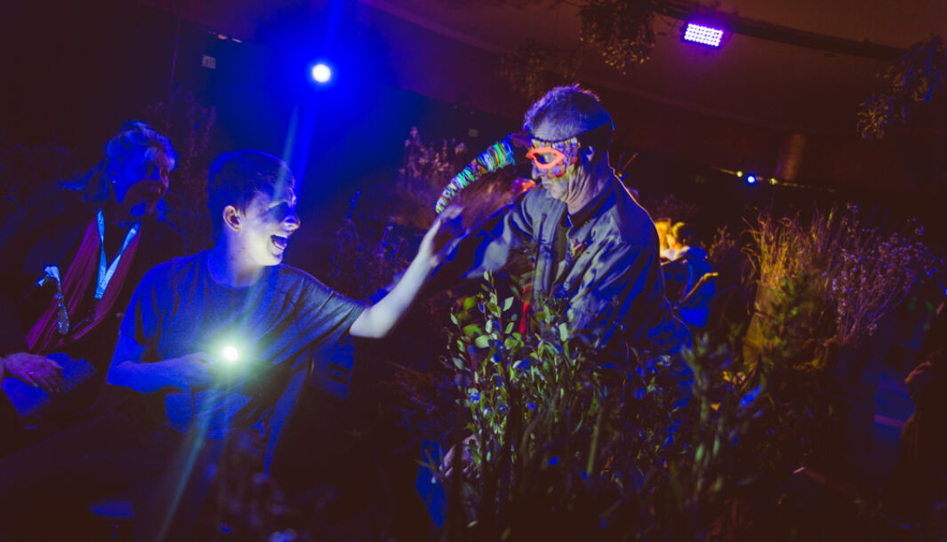 A When the World Turns production photo. A Polyglot artist in an intricate mask leans over a bank of plants towards a young person, who holds a small torch, and reaches towards them smiling. An adult watches the exchange, smiling. They are in a darkened space, illuminated with dark blue theatrical lighting. Photographer: Theresa Harrison