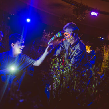 A When the World Turns production photo. A Polyglot artist in an intricate mask leans over a bank of plants towards a young person, who holds a small torch, and reaches towards them smiling. An adult watches the exchange, smiling. They are in a darkened space, illuminated with dark blue theatrical lighting. Photographer: Theresa Harrison
