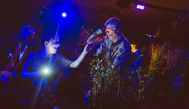 A When the World Turns production photo. A Polyglot artist in an intricate mask leans over a bank of plants towards a young person, who holds a small torch, and reaches towards them smiling. An adult watches the exchange, smiling. They are in a darkened space, illuminated with dark blue theatrical lighting. Photographer: Theresa Harrison
