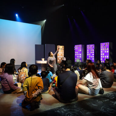 A Come Back Home production photo. A group of children and adults sit on the floor under theatre lighting. They face a performer who points to a dimly-lit ceiling. The audience is surrounded by cardboard silhouettes of skyscraper buildings. Photo: Studio Znke, courtesy of Esplanade - Theatres on the Bay, Singapore.