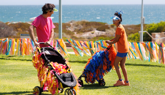 A Pram People production photo. Two performers stand on a grass lawn pushing prams. The prams are decorated with colourful streamers. In the background is the ocean. Photo: Travis Hayto from SoCo Studios.