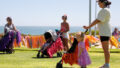 A Pram People production photo. Three parents stand on a grass lawn with prams. The prams are decorated with colourful streamers. In the background is the ocean. Photo: Travis Hayto from SoCo Studios.