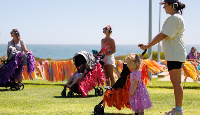 A Pram People production photo. Three parents stand on a grass lawn with prams. The prams are decorated with colourful streamers. In the background is the ocean. Photo: Travis Hayto from SoCo Studios.
